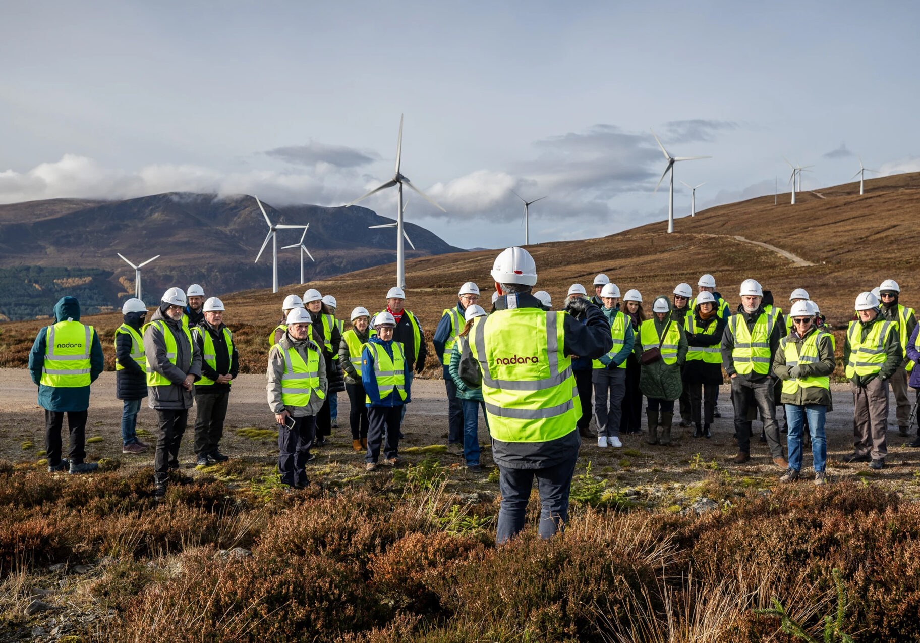 Nadara team in hi-vis vests and safety helmets, at a windfarm site with turbines in the background and heather underfoot.