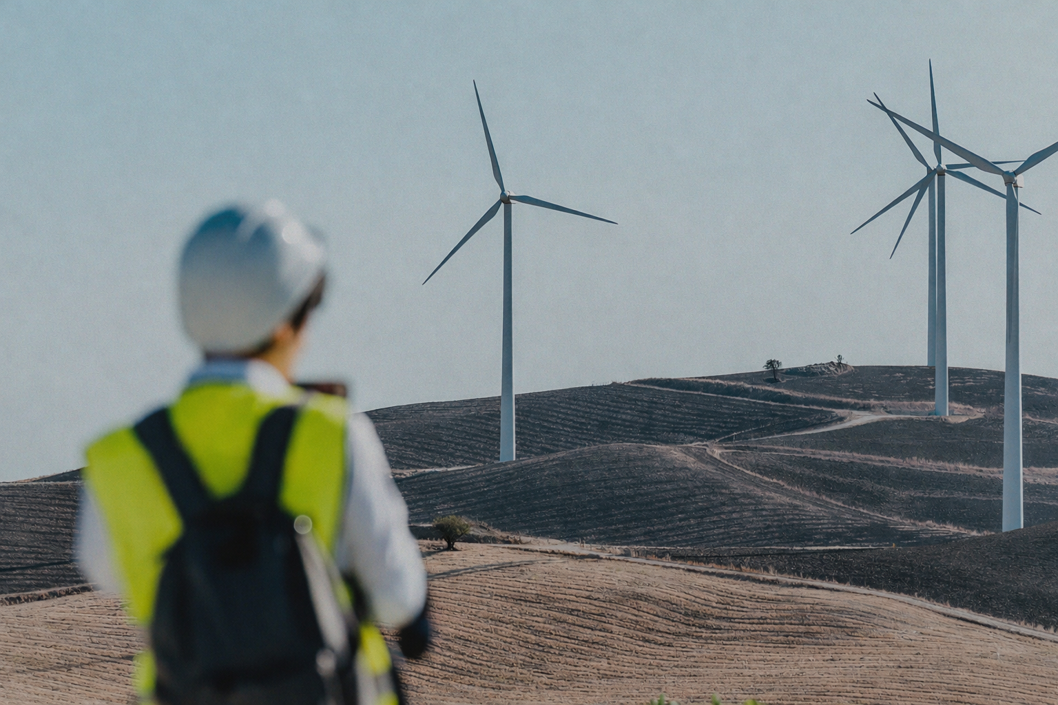 A technician in a hard hat and hi-vis vest looks towards wind turbines in the distance